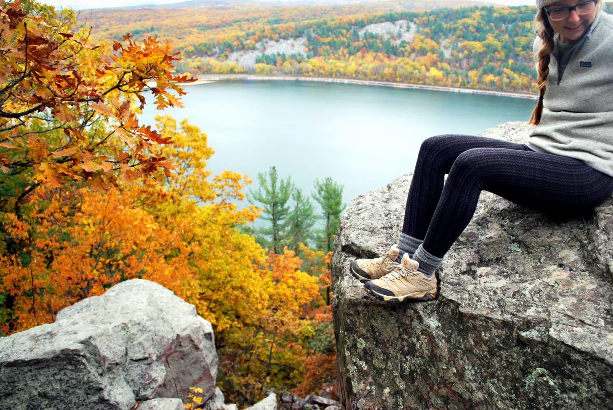 Woman sitting on rock cliff above blue lake surrounded by a forest of trees with orange and yellow leaves.
