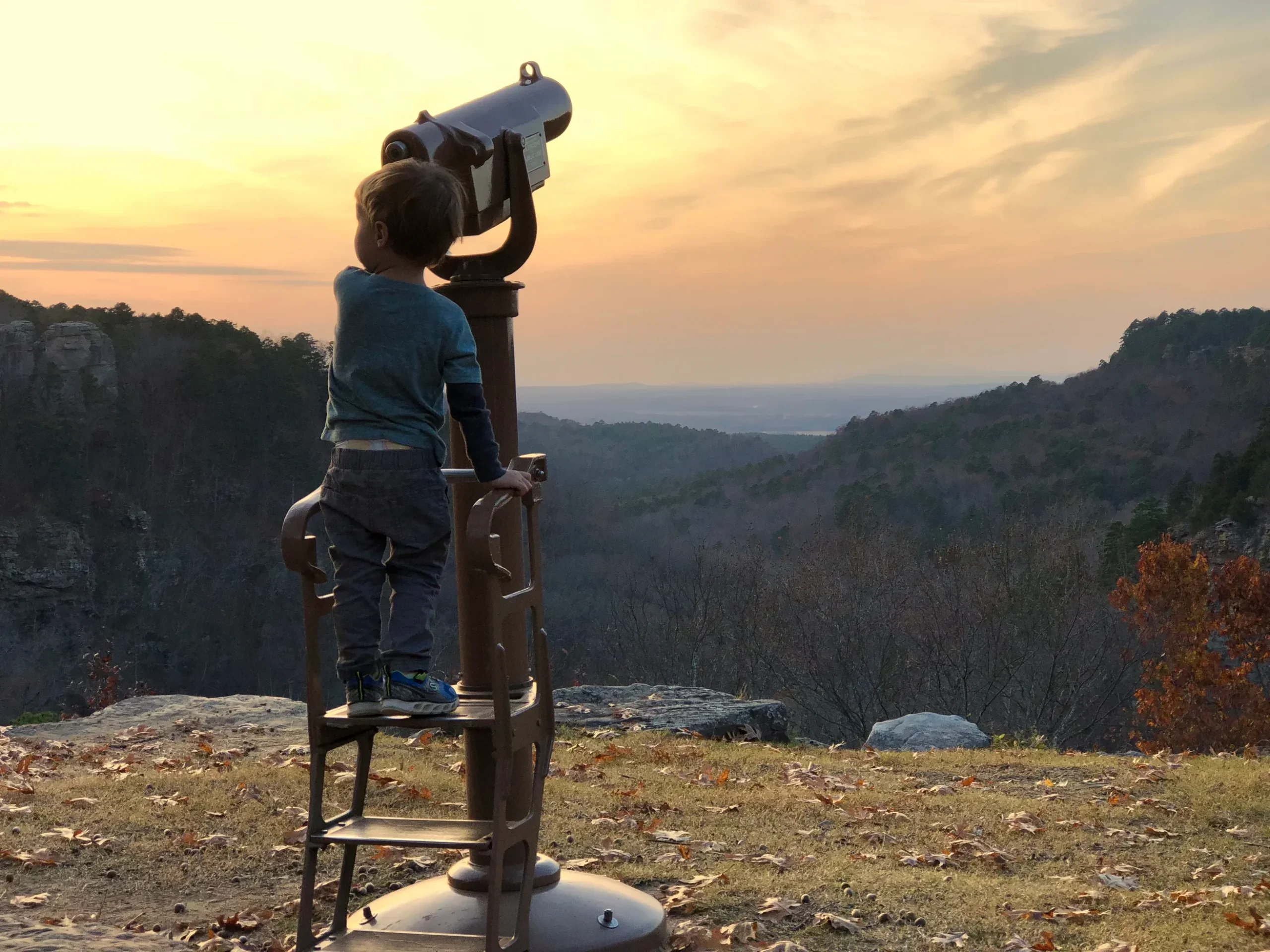 Toddler standing at a statinary telescope at an overlook during sunset.