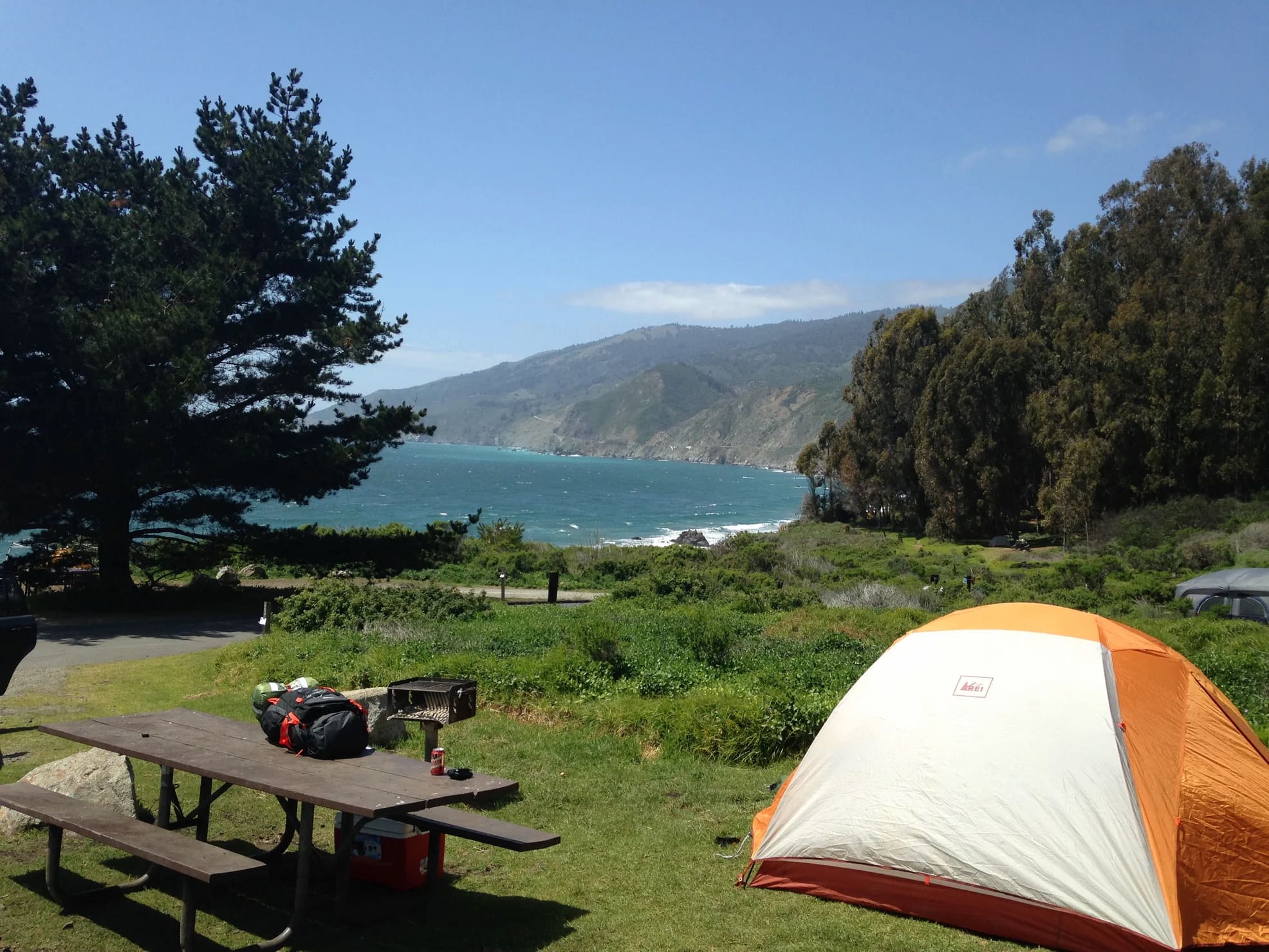 Orange tent at grass covered campsite with picnic table overlooking the Pacific coast and blue ocean down below.