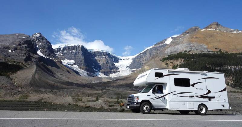 Class C RV driving up a mountain road with snow-covered mountains behind.