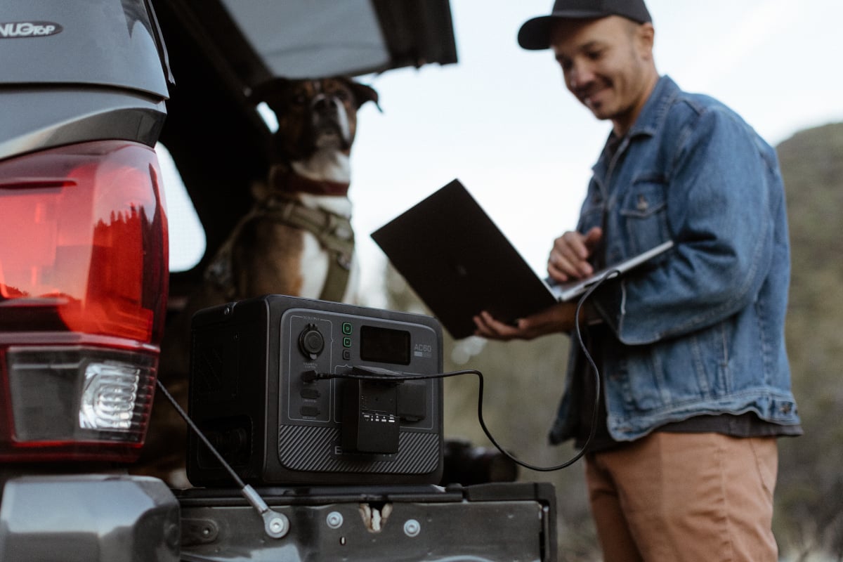BLUETTI AC60 set on the tailgate of a vehicle with dog and man in the background. The man has his laptop plugged into the power station.