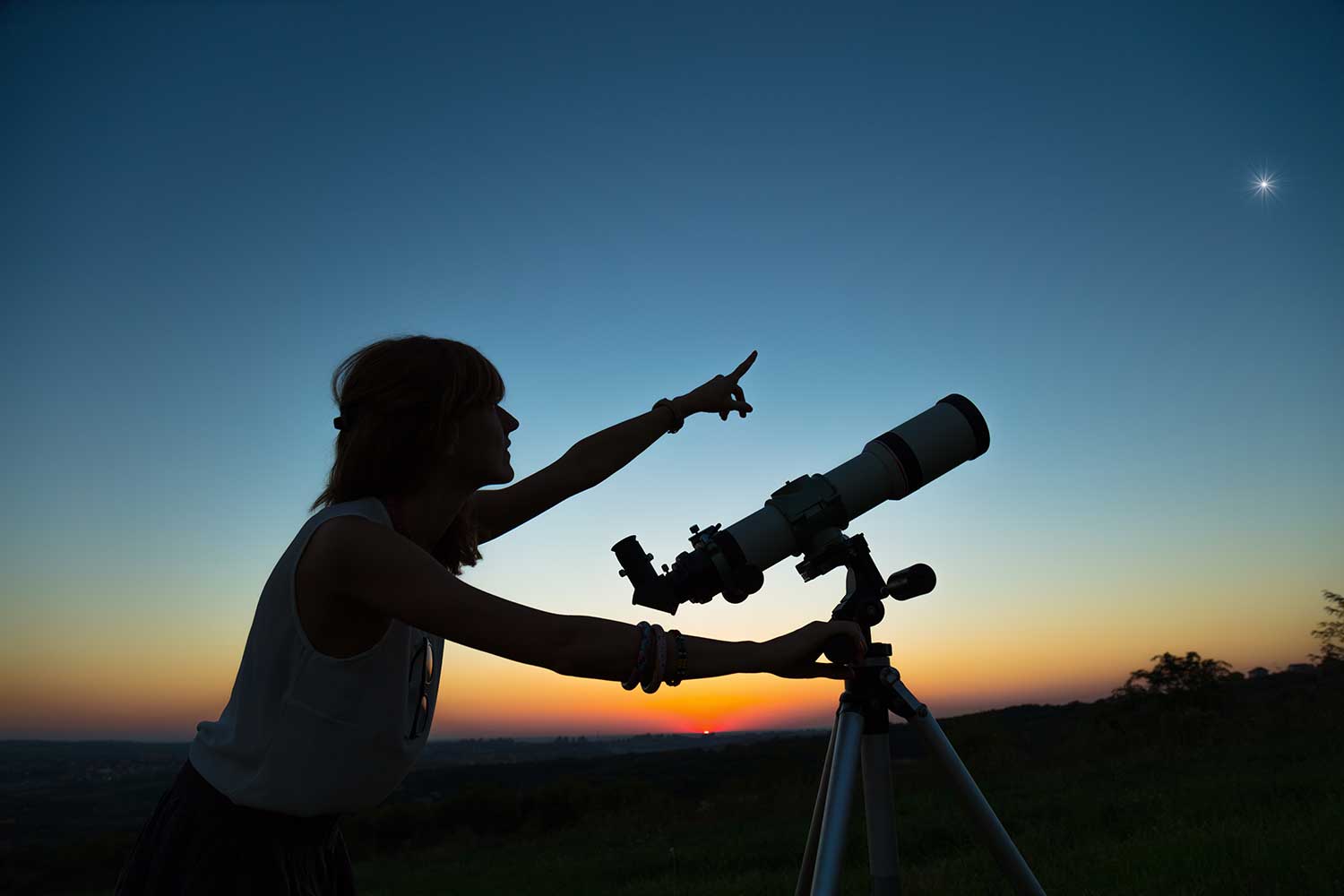 womans stands near telescope and points at a dark blue sky 