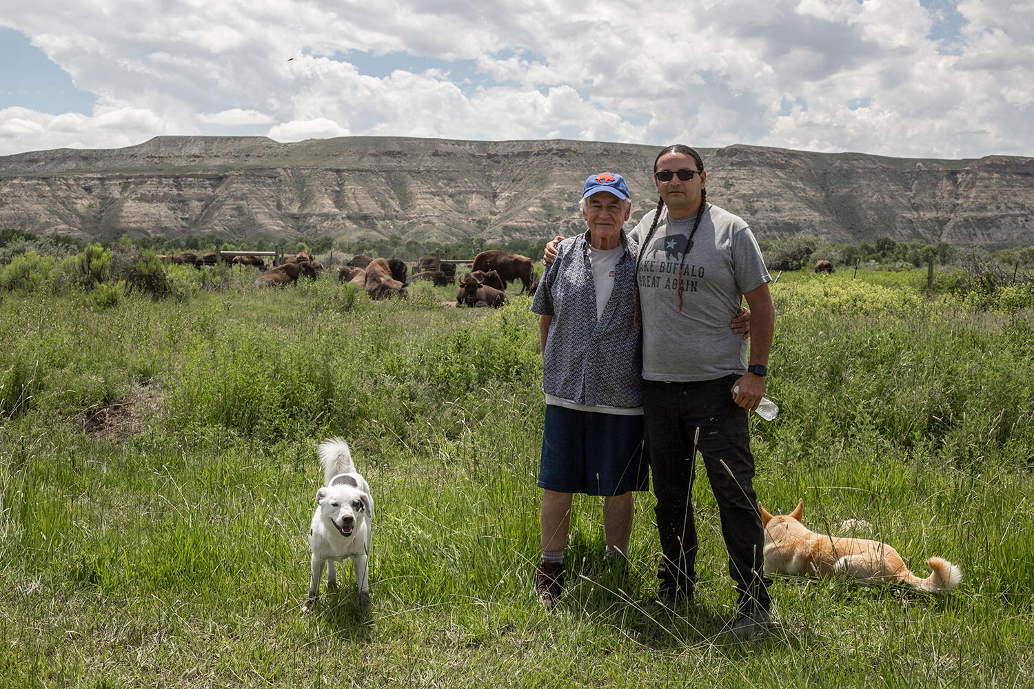 two men and stand on prairie with two dogs; buffalo and cliffs behind them