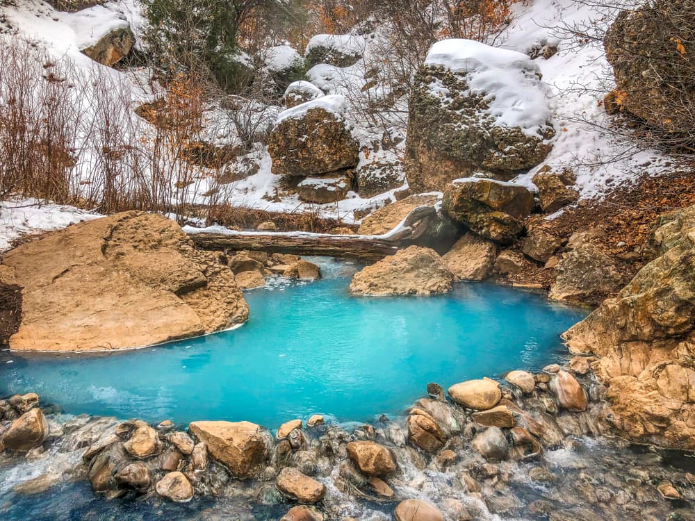 bright teal hot springs surrounded by craggy brown rocks, lightly covered in snow