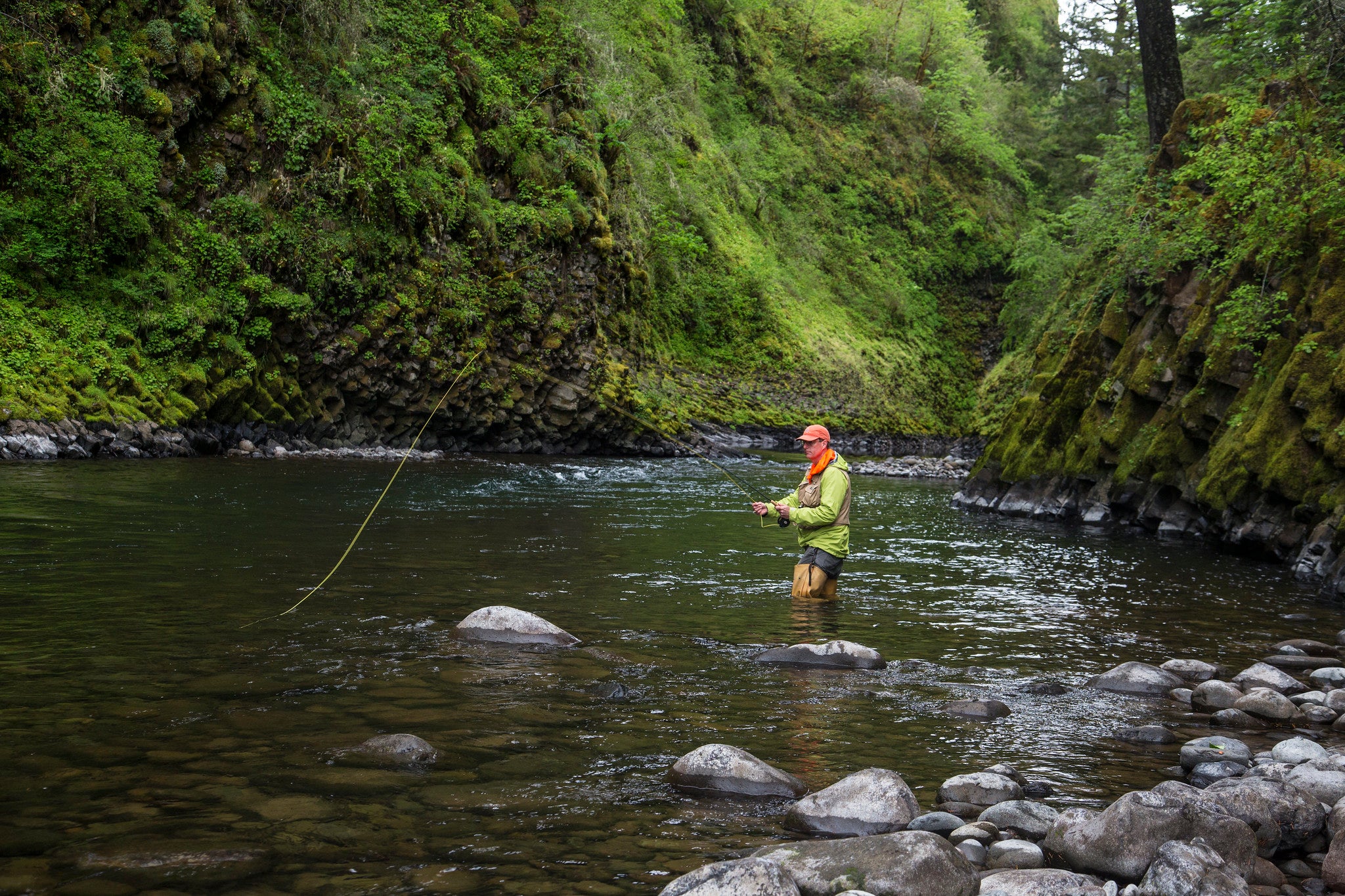 A man in a greenshirt and waders fly fishes in a shady river.
