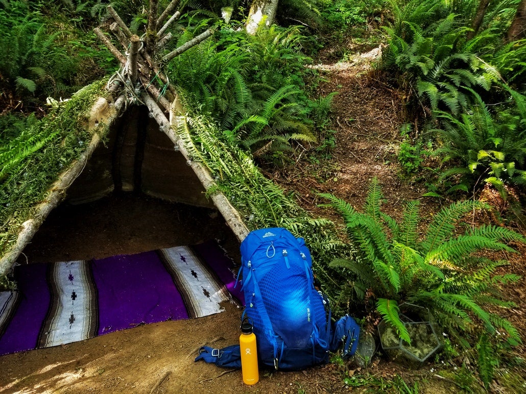 A primitive tent made from grass and sticks in the woods, with a backpack out front. backpacks are found on our primitive camping checklist