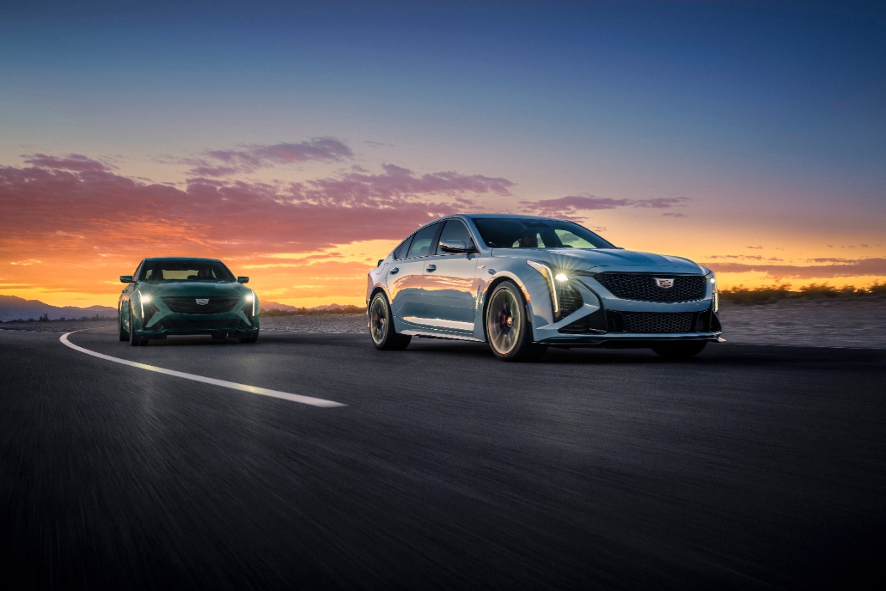Front passenger view of the 2025 Cadillac CT5-V in Typhoon Metallic (left) and CT5-V Blackwing in Drift Metallic (right) driving at dusk.