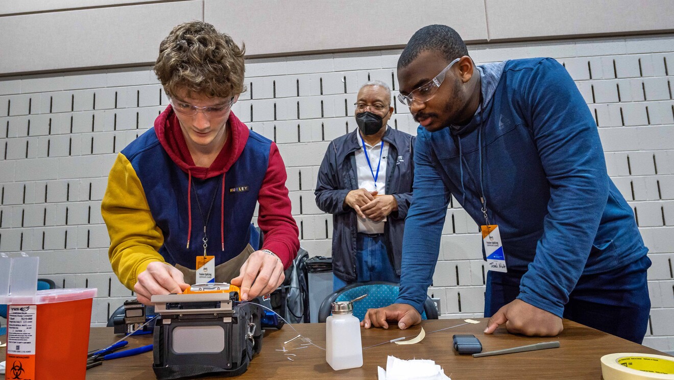 A photo of two students working on a fiber optic fusion splicer. An instructor is standing behind them.