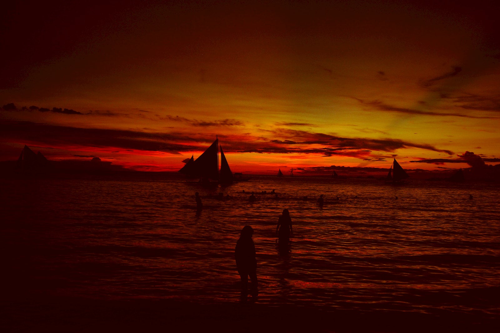 People on the beach overlooking sailboats at sunset.