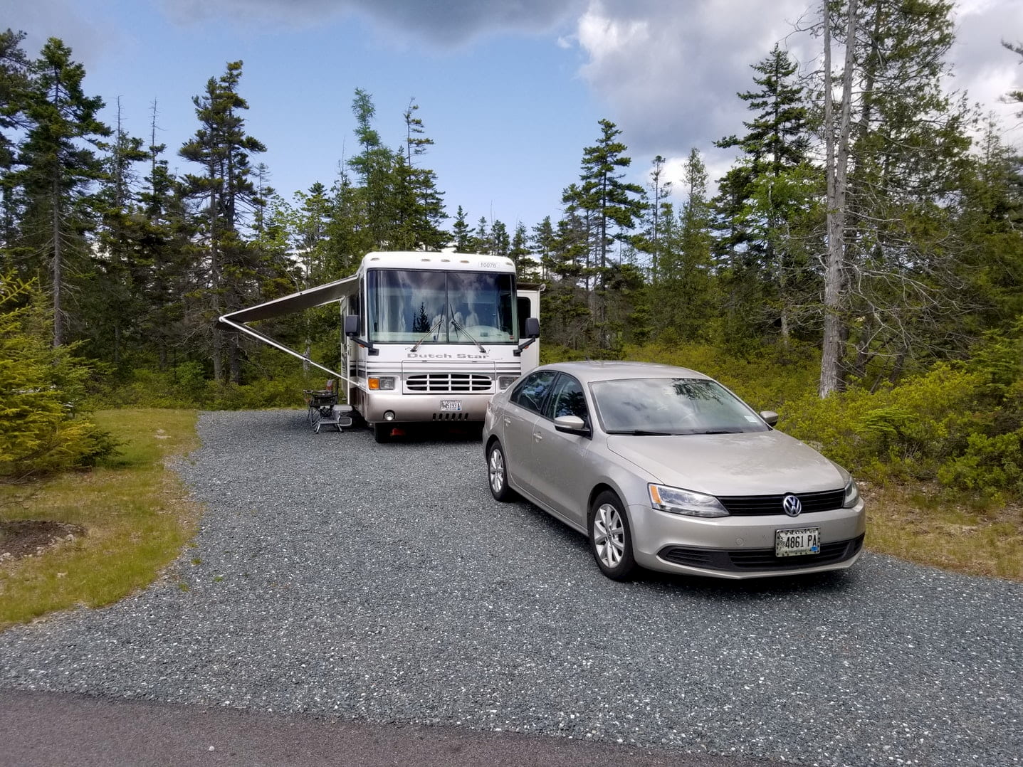 R.V. and car parked in gravel campsite beside pine trees.