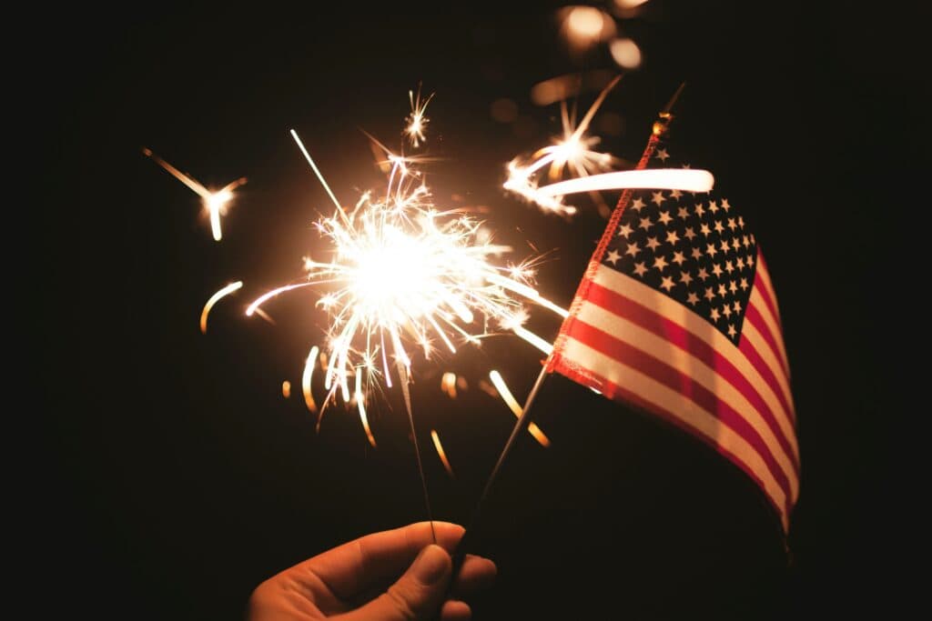 A hand holding a lit sparkler and waving a small American flag during a Fourth of July fireworks display.