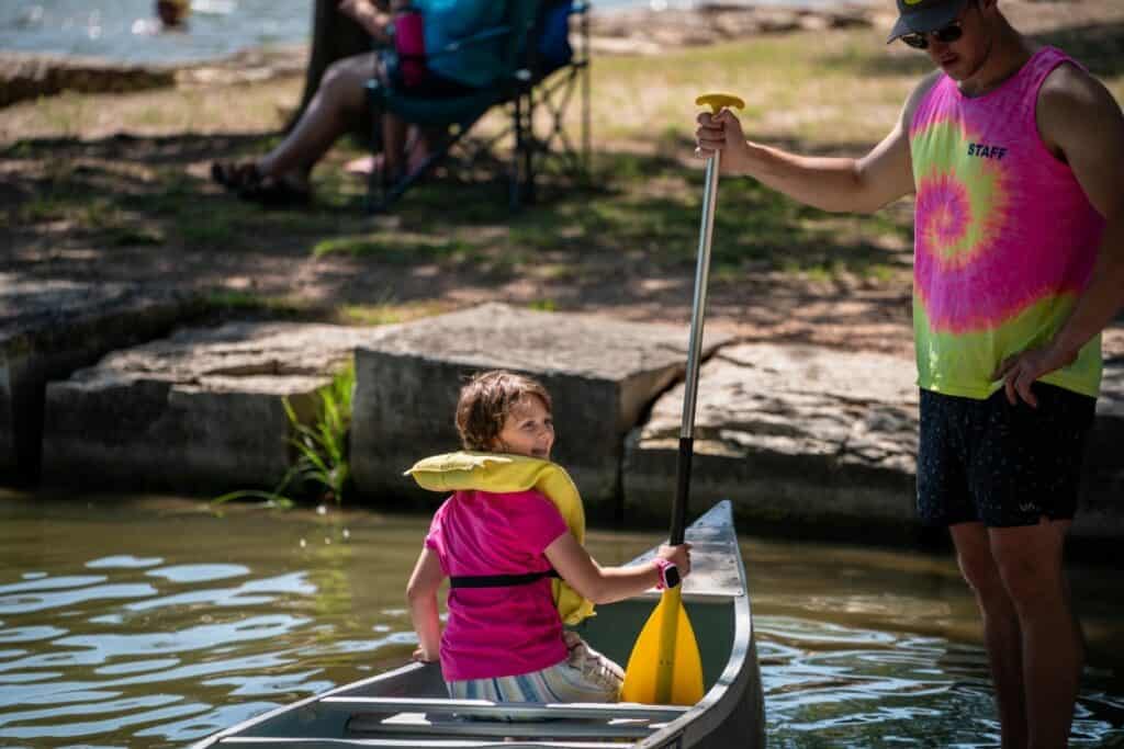 boy paddles canoe in river with staff watching over.