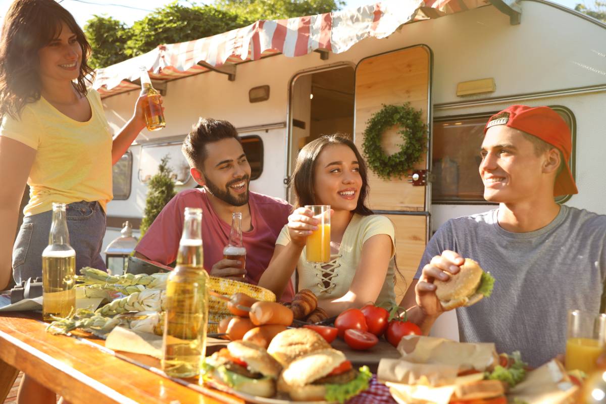 Happy RVing friends with drinks and food at table near travel trailer. 