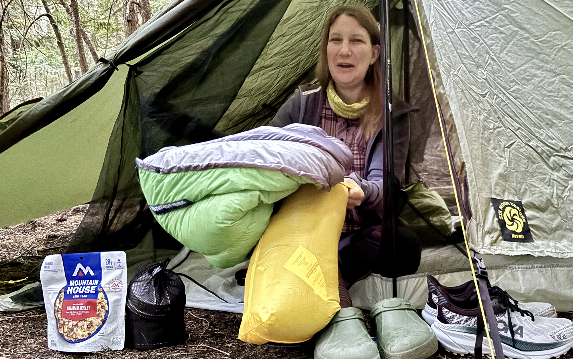 a woman stuffs a sleeping bag into a stuff sack while sitting inside a tent in the morning