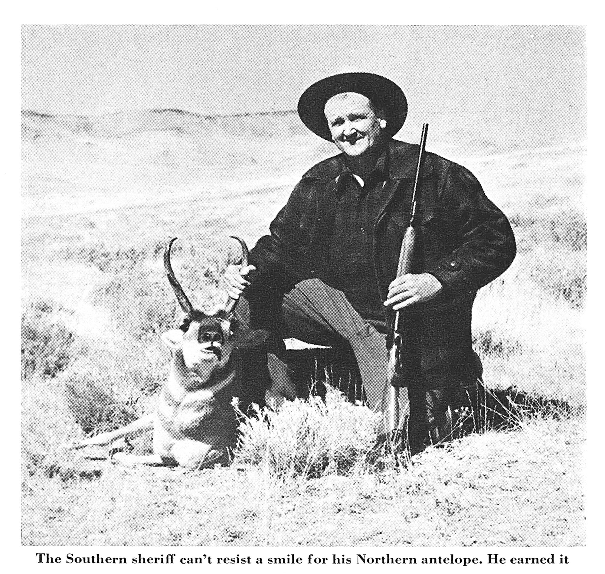 A hunter in a black shirt and hat kneels beside a buck pronghorn in a black and white photo.