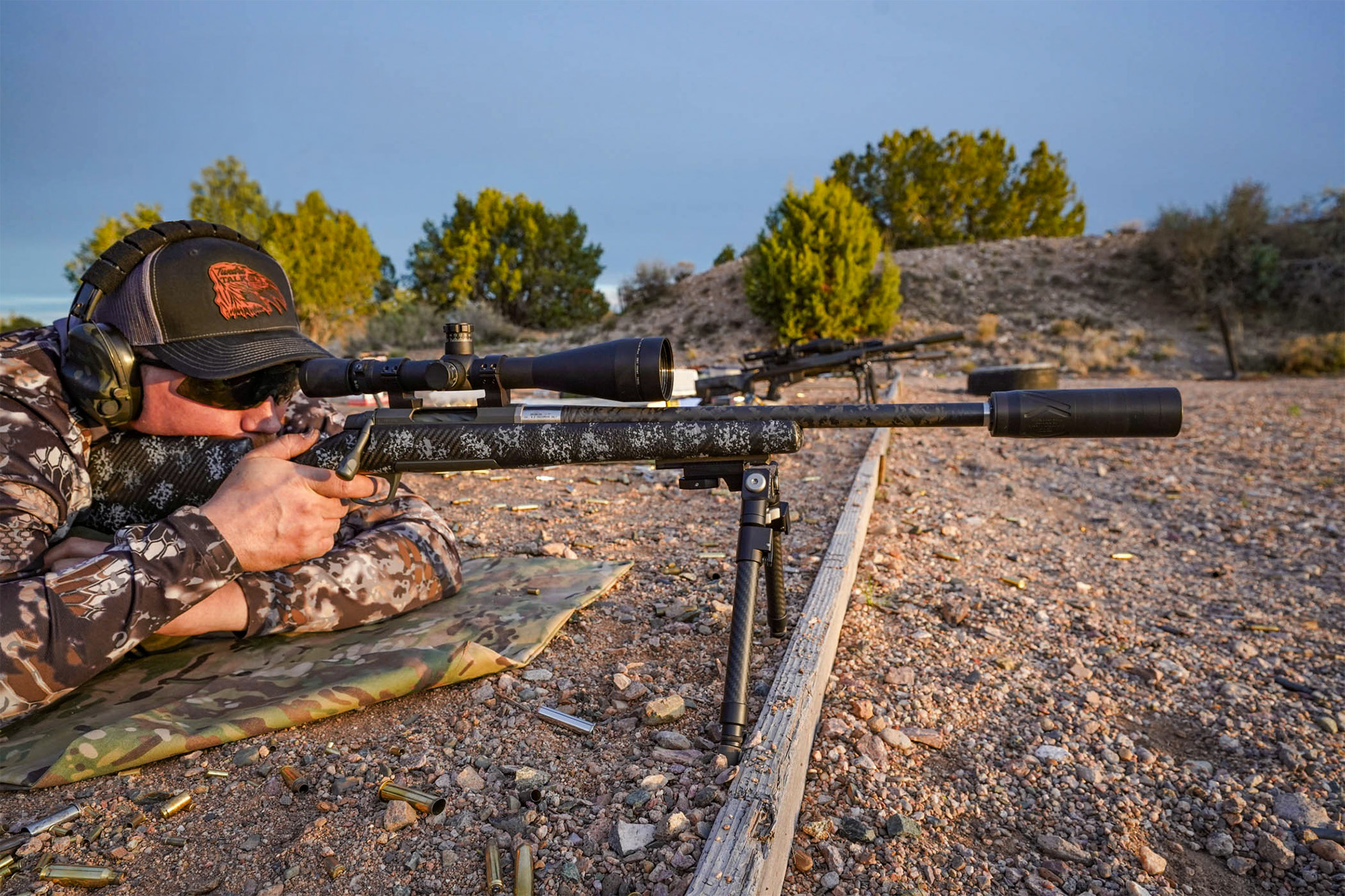 A man lies prone at the range, behind a rifle with a suppressor on it.