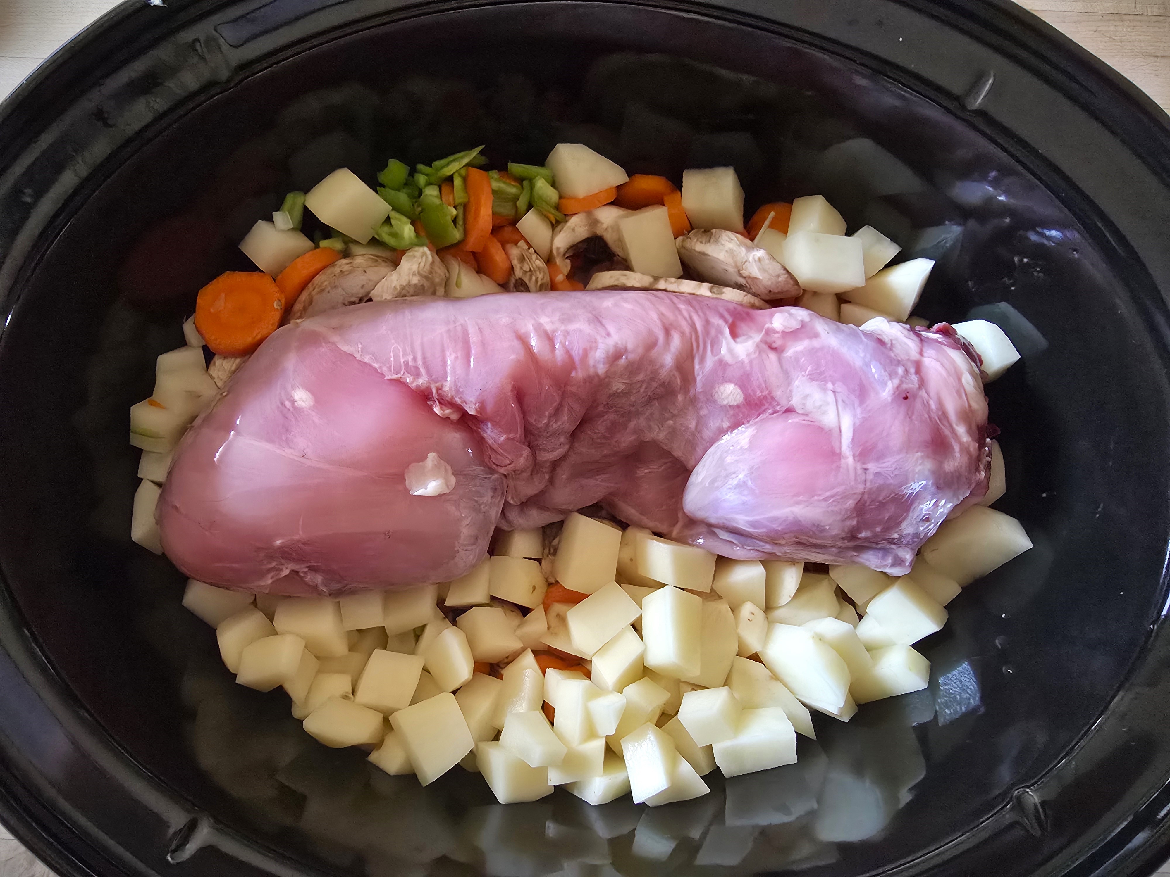 A skinned rabbit in a crockpot surrounded by chopped vegetables.