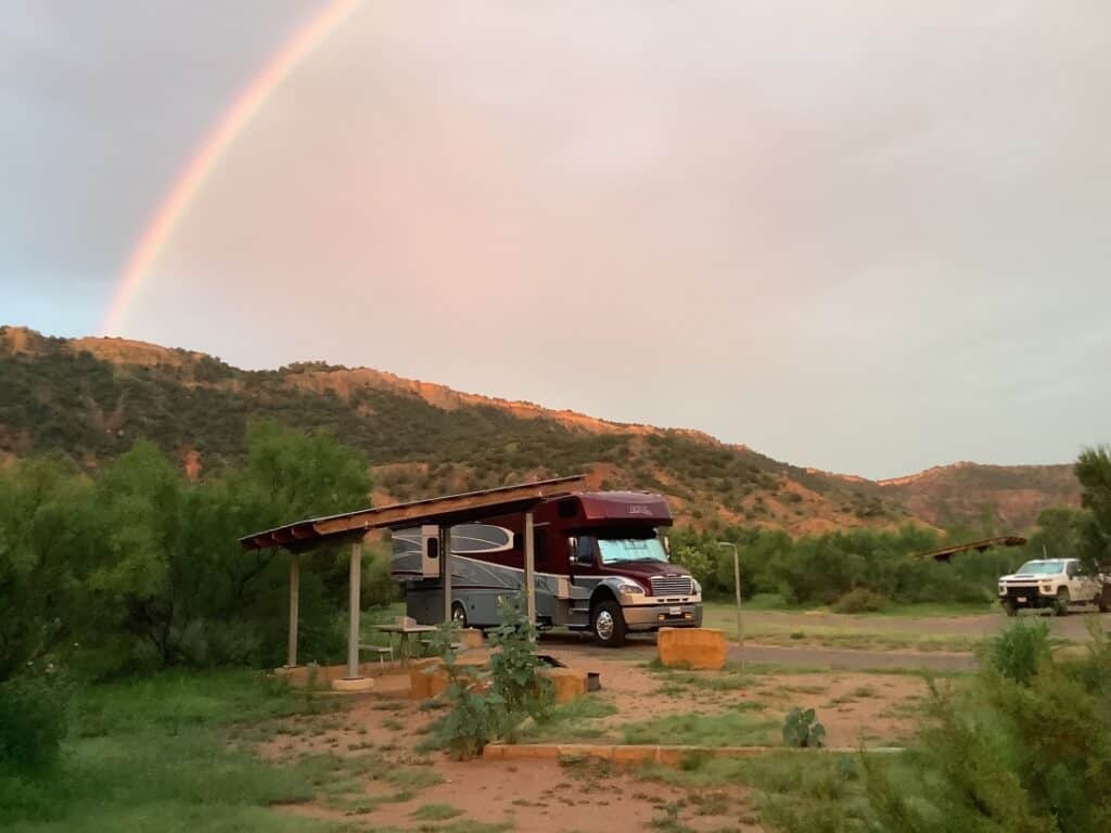 A large Class C motorhome in a campsite in Palo Duro SP with a rainbow overhead.