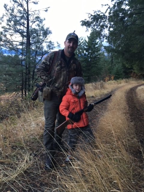 A man and a young boy cross a dirt path carrying rifles. 