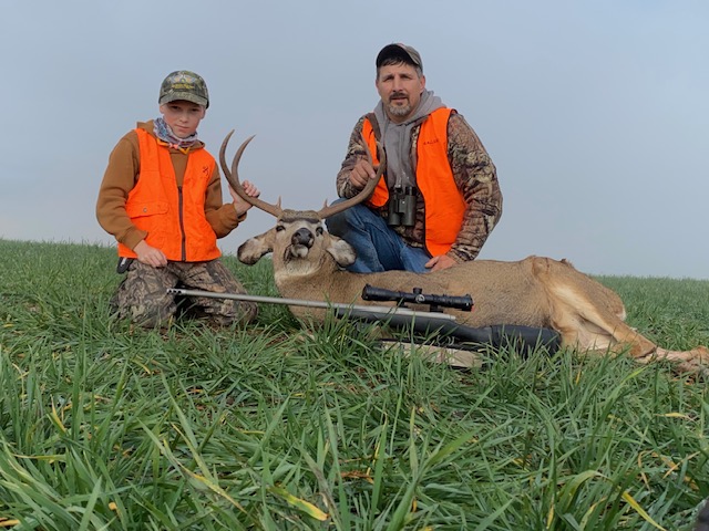 Two hunters in orange vests pose with a mule deer. 