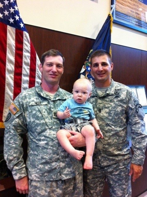 Two soldiers in Army uniforms pose with a young baby. 