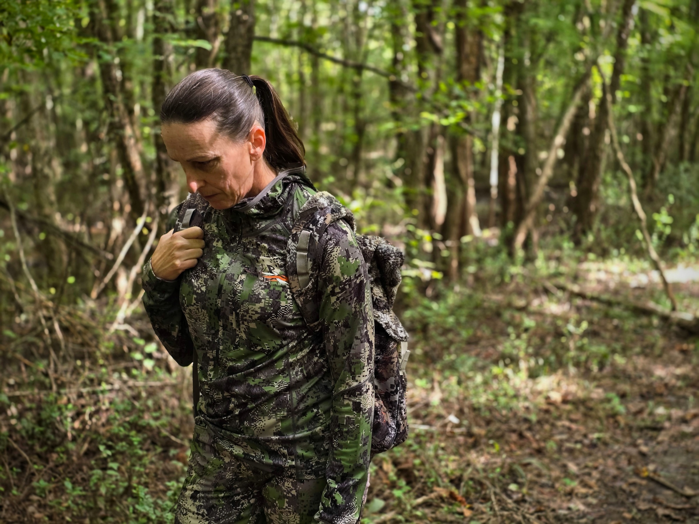 A woman in green camouflage walks through the woods carrying a backpack.