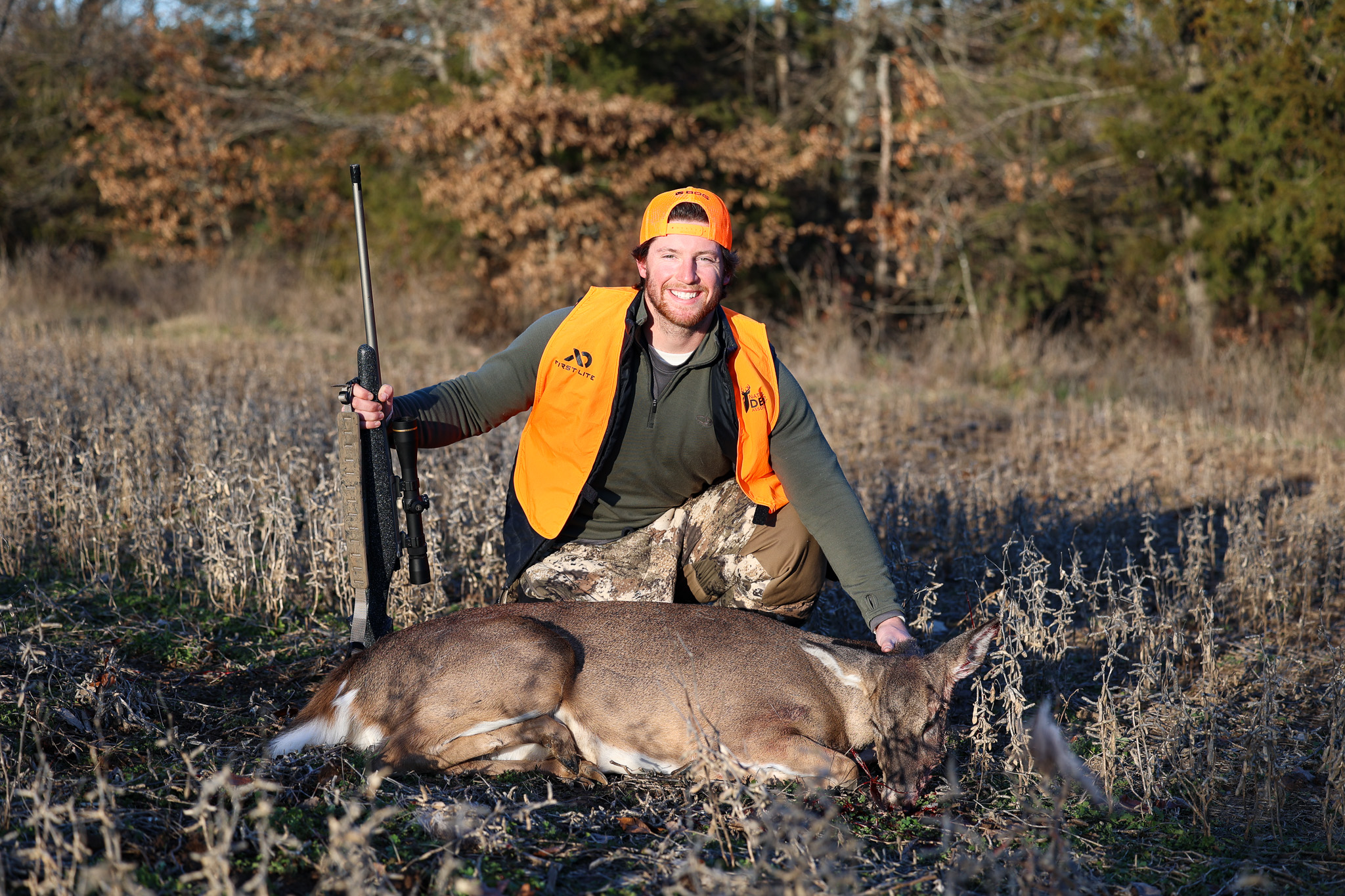 A man in blaze orange and a backward hat kneels beside a doe in a bean field.