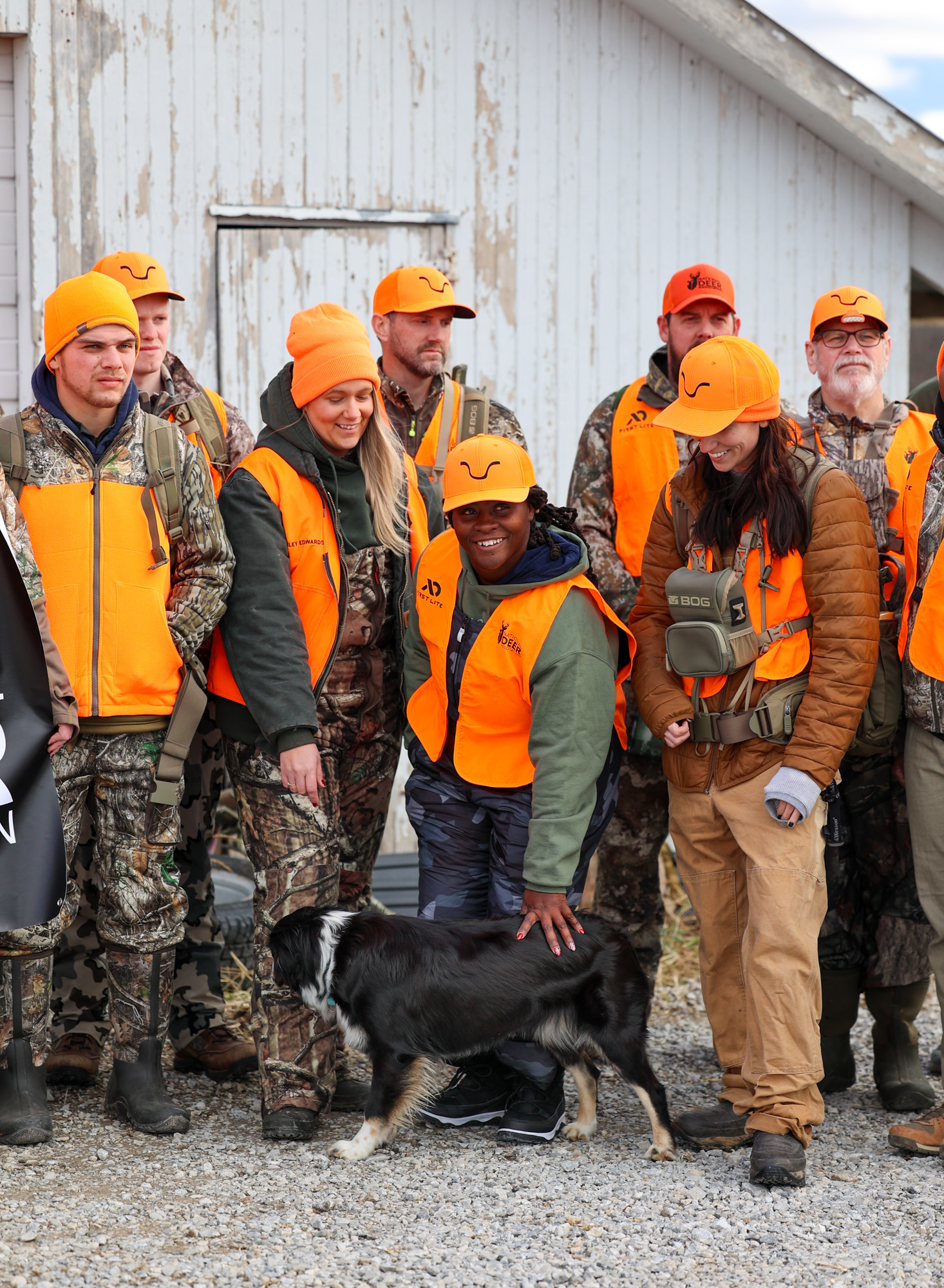 A group of hunters in blaze orange laugh and pet a dog.