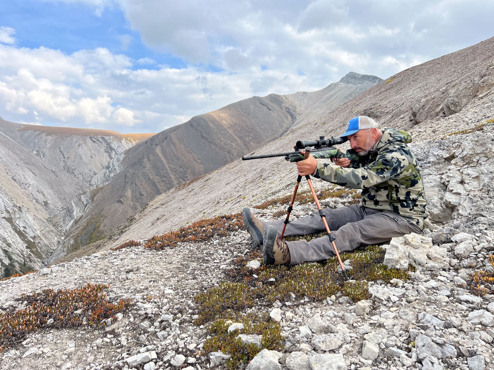 A man sitting on a rocky landscape aims a rifle balanced on shooting sticks