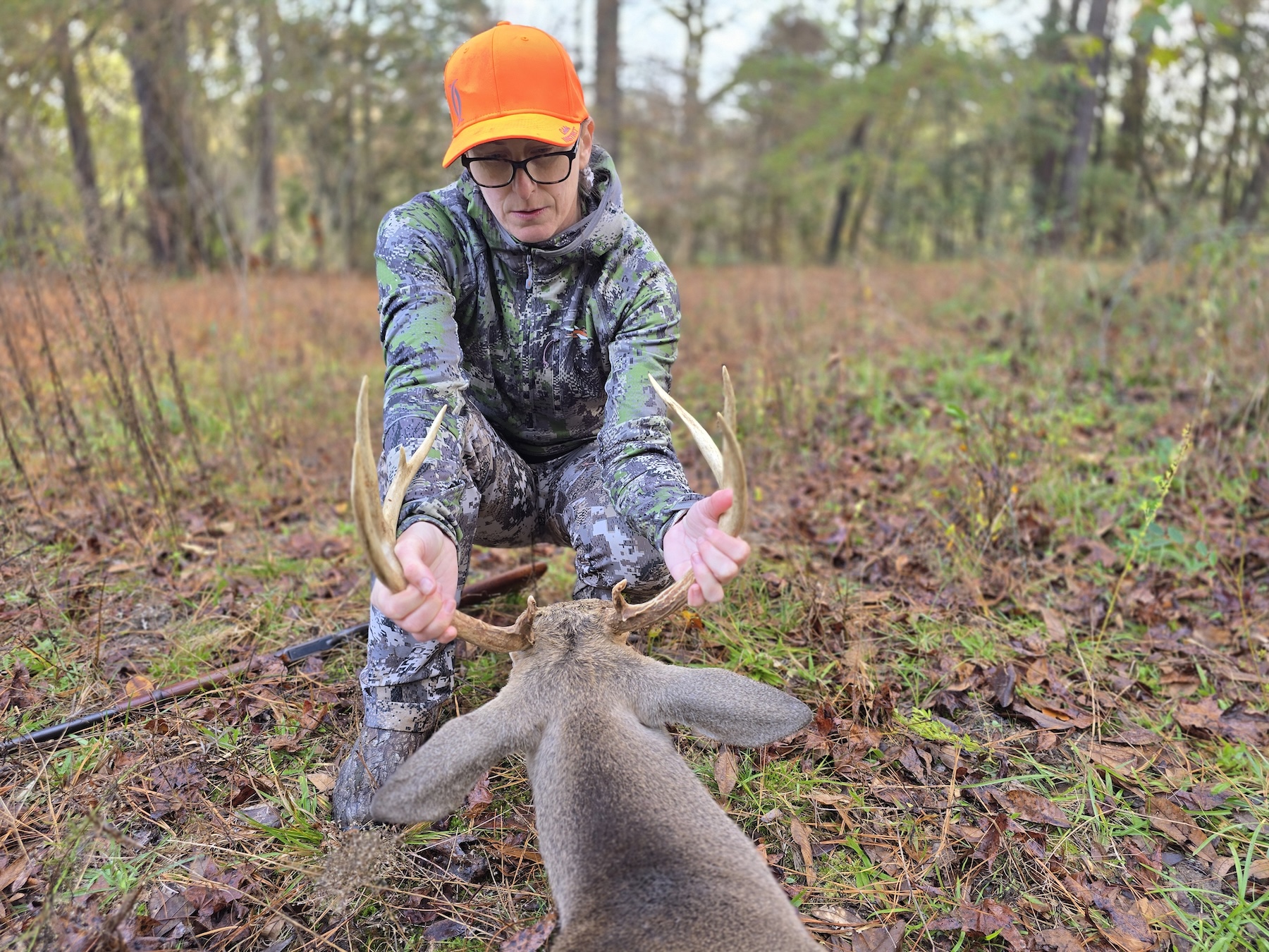 A woman wearing camo and an orange hat holds the rack of a whitetail buck. 