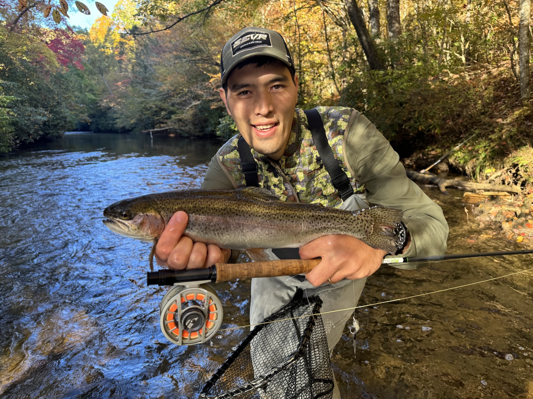 A man in a hat holds a rainbow trout and a fly rod. 