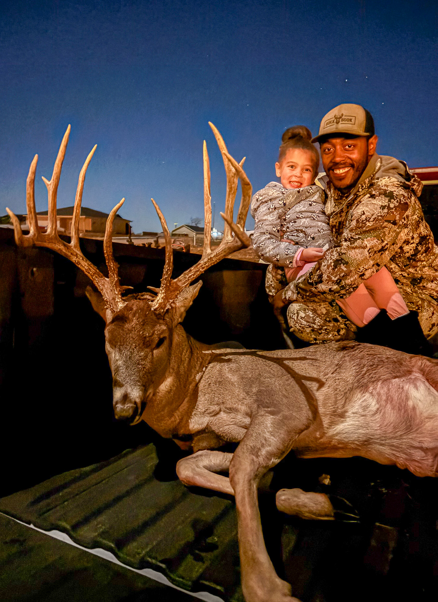 A bowhunter and his daughter smile beside a big buck in a truck.