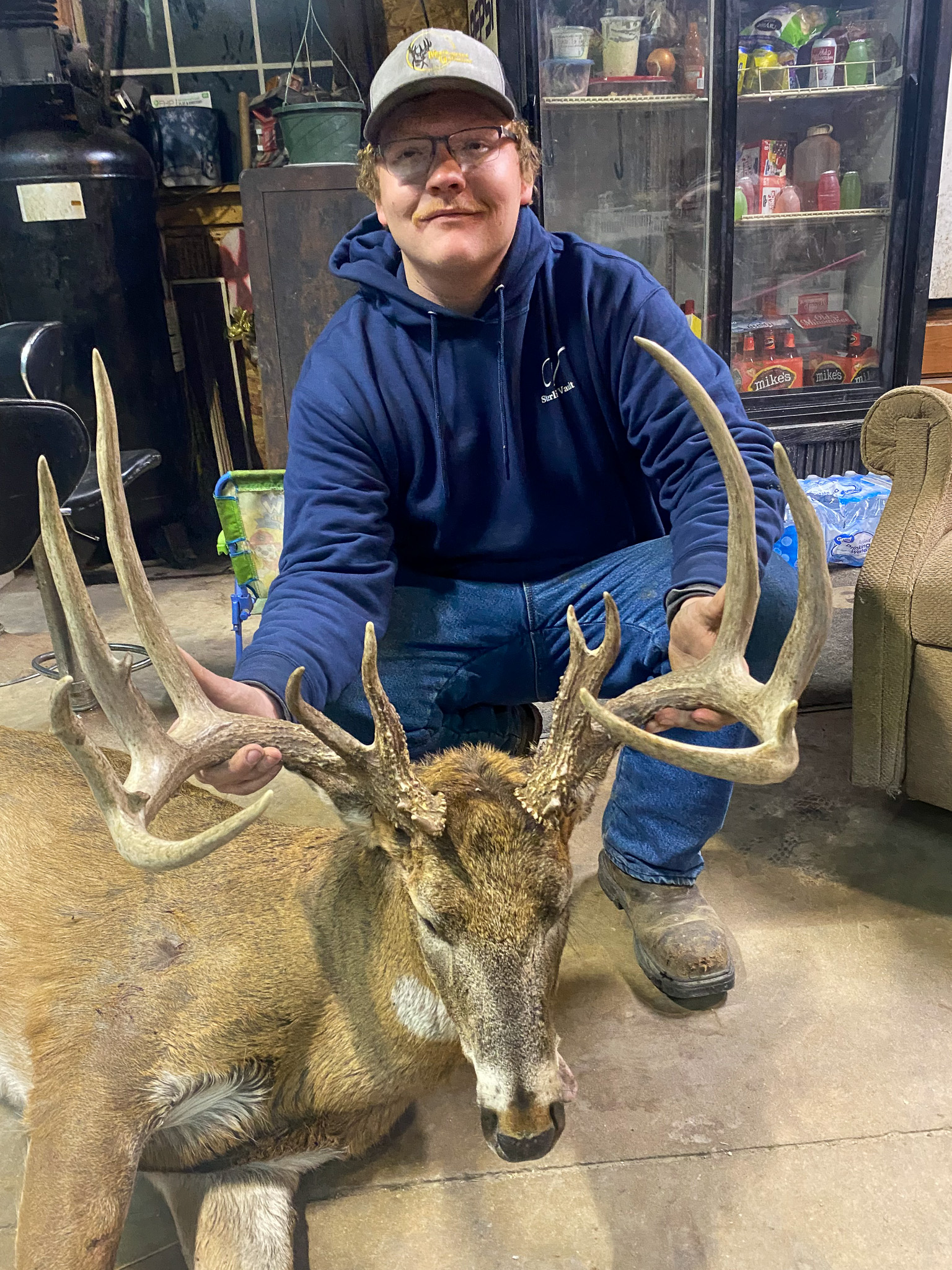 A hunter in a blue hoodie kneels behind a split-brow-tine buck.