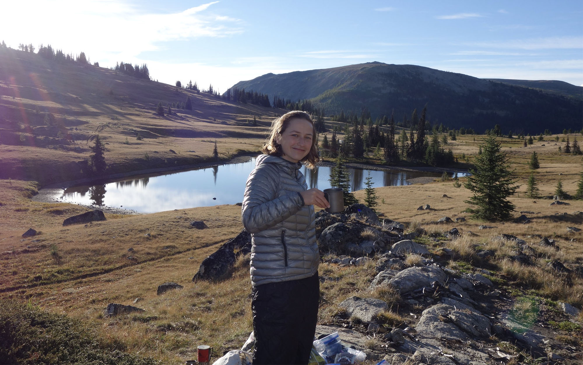 woman with a coffee mug smiling with a lake and mountain scene in the background