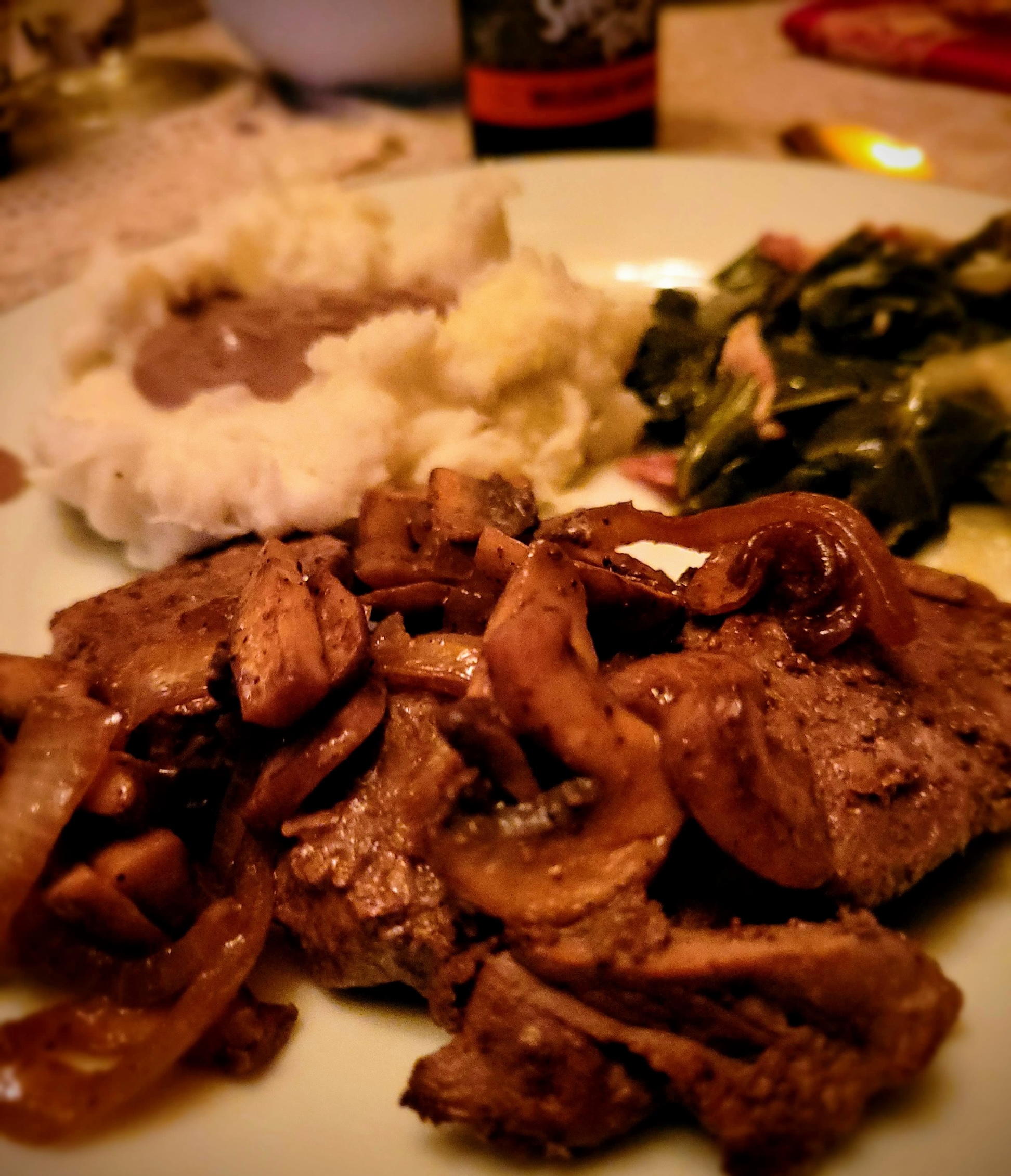 A plate of venison backstrap steaks topped with grilled mushrooms and onions and served collards, mashed potatoes, and gravy.