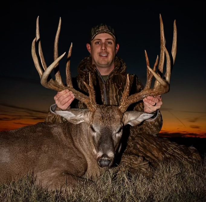 A man holds up the rack of a large 14 point whitetail buck as the sun sets behind him.