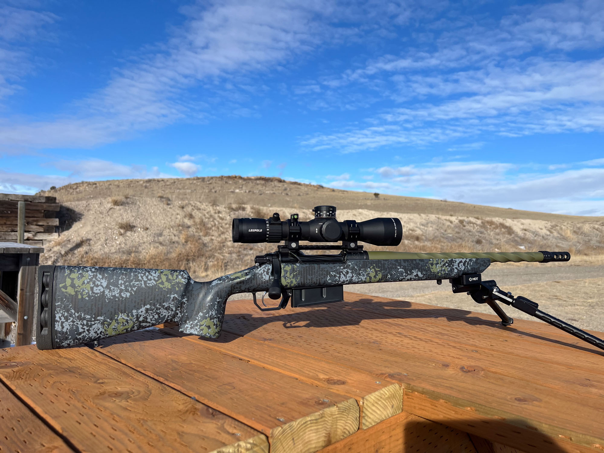 A camo rifle with a short fluted barrel and muzzle brake resting on a shooting bench. The rifle is chambered in 7mm backcountry.