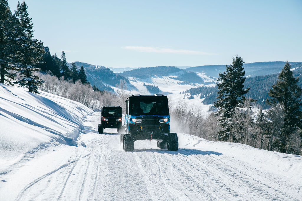 Two Can-Am side-by-sides with tracks drive on a snowy mountain trail.