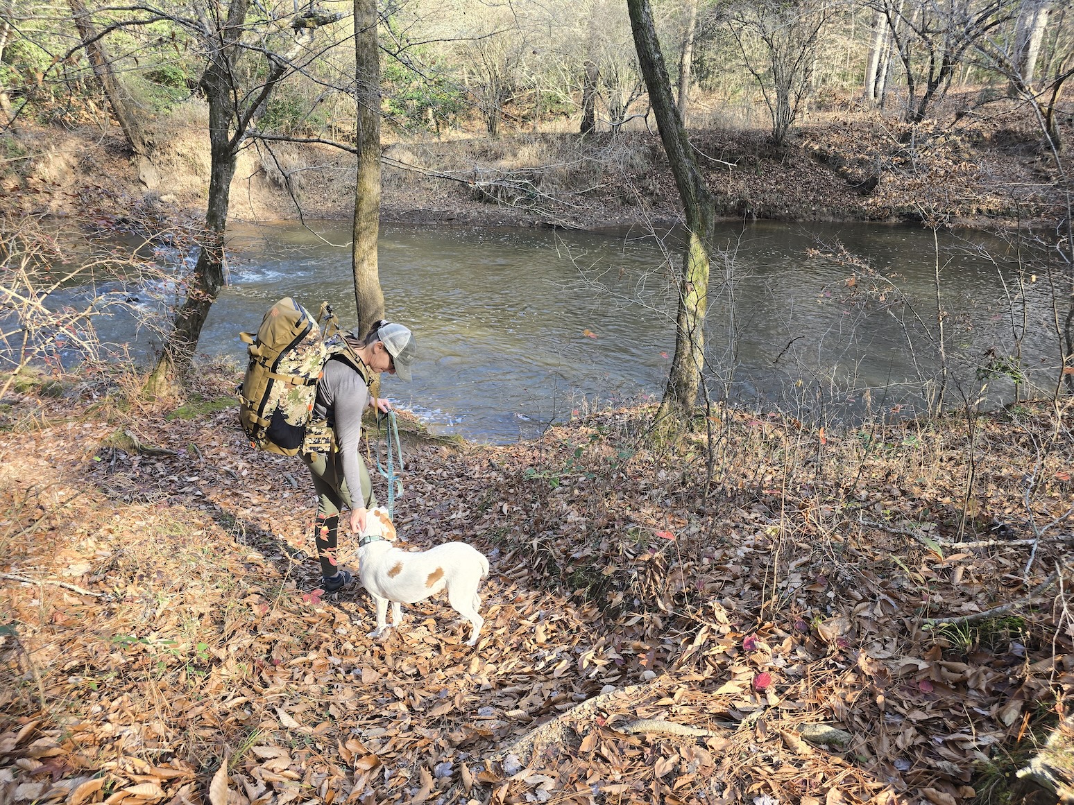 A woman carrying a loaded backpack pets a white dog on a leash near a creek.