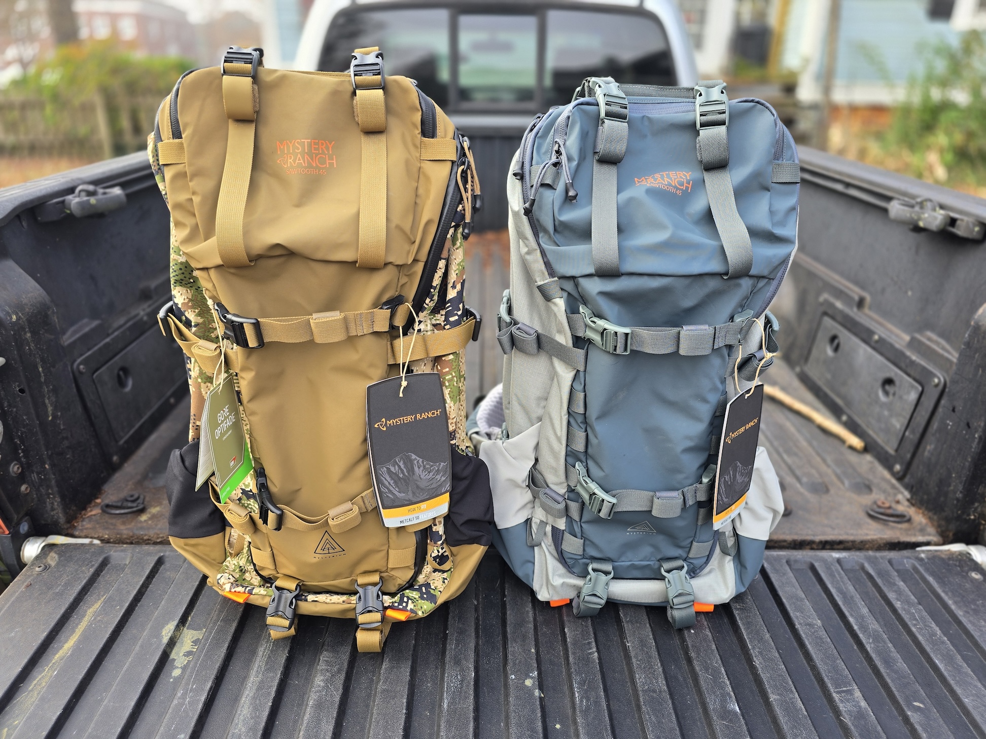 Two Mystery Ranch backpacks with tags sit on the tailgate of a pickup truck.