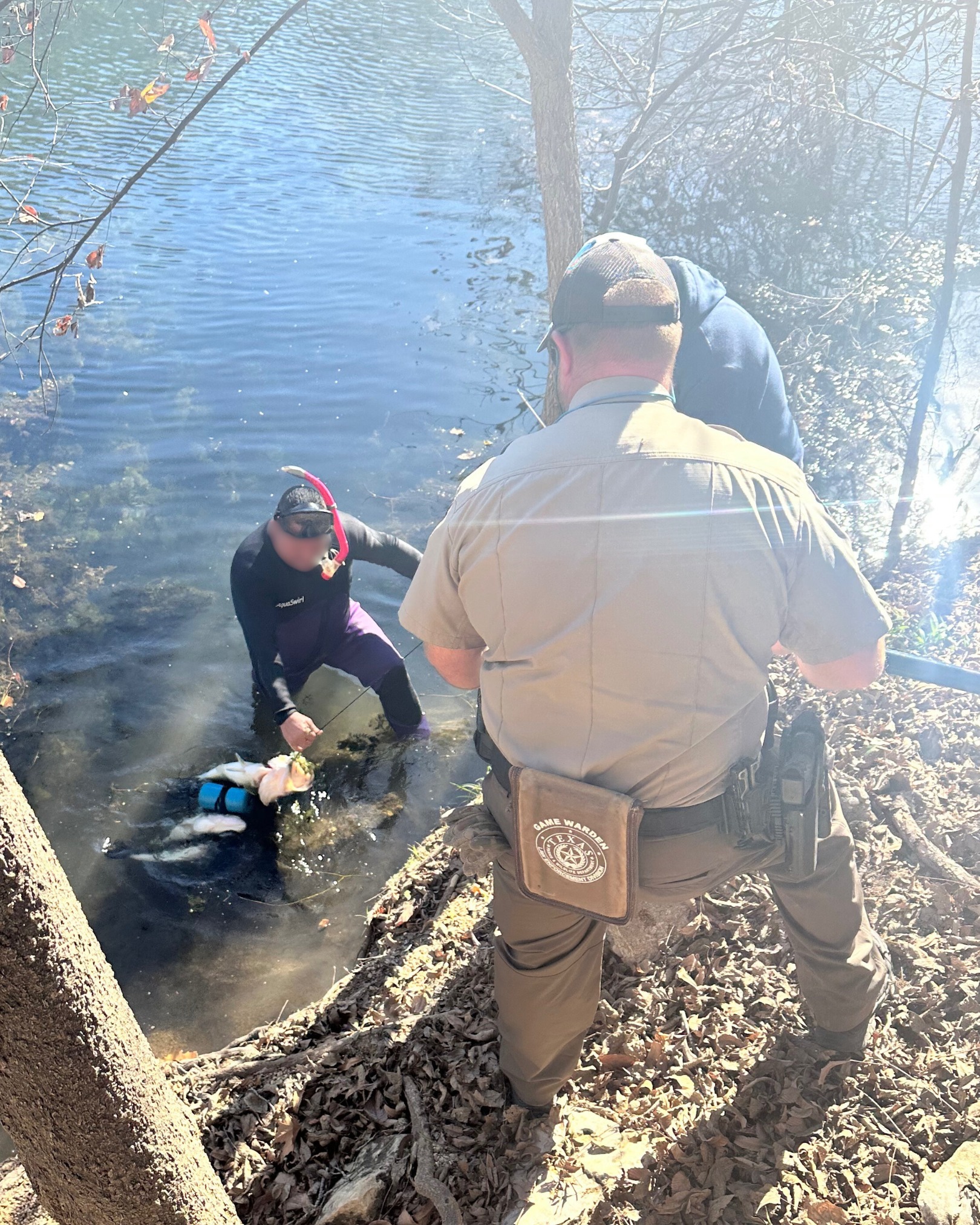 A game warden in uniform waits on the bank while a man in a wet suit and snorkel climbs ashore with a speared largemouth bass.