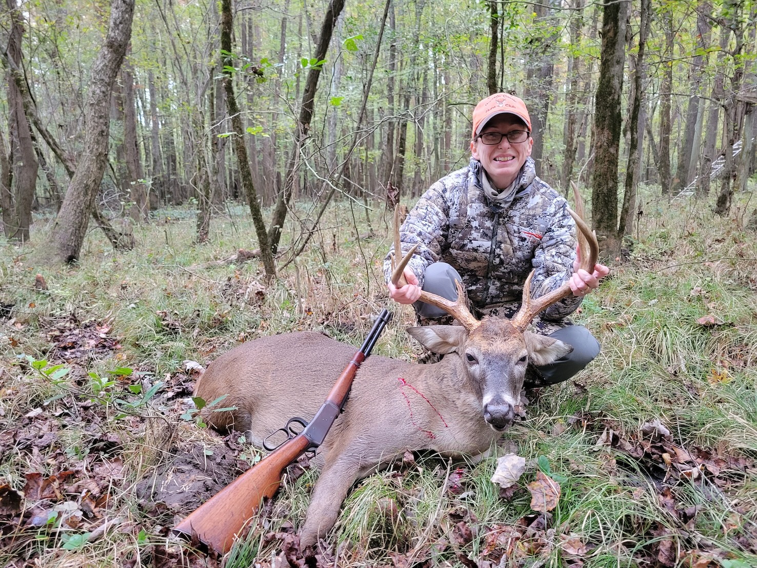 A woman in camo and an orange hat holds up the rack of a large 8-point buck with a lever-action Winchester Model 94 laid against the deer.