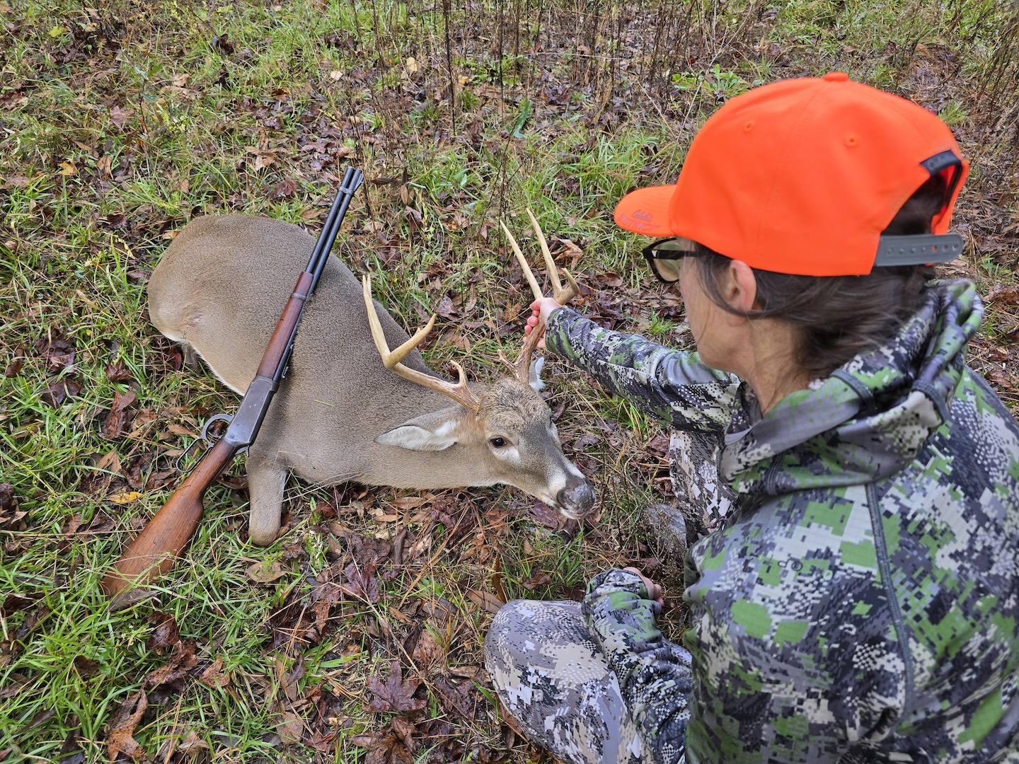 A woman dressed in camo with a blaze orange hat looks at the rack of a 10-point buck. There is a Winchester Model 94 laid against the deer's side.