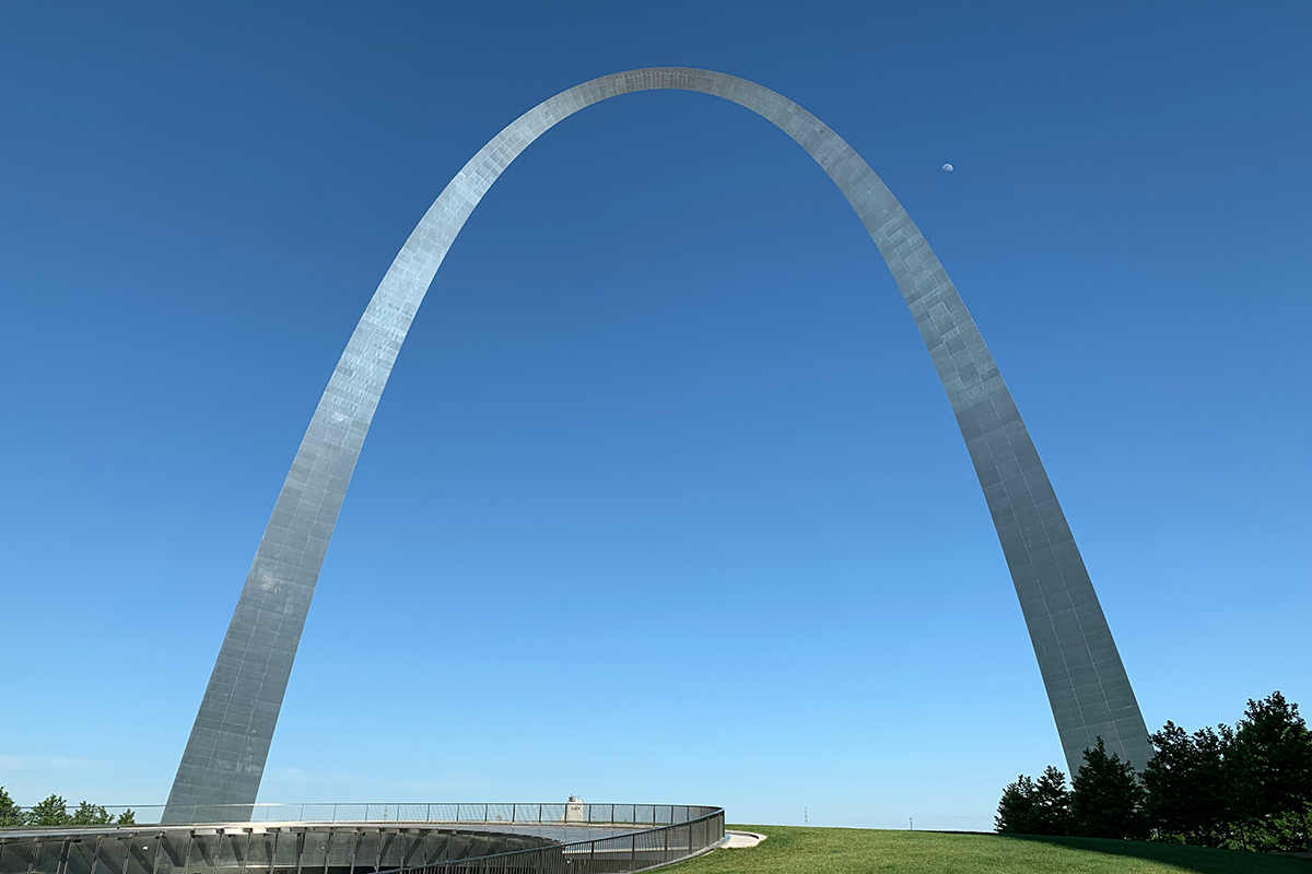 Graceful steel monument arcs over a green field against a blue sky.