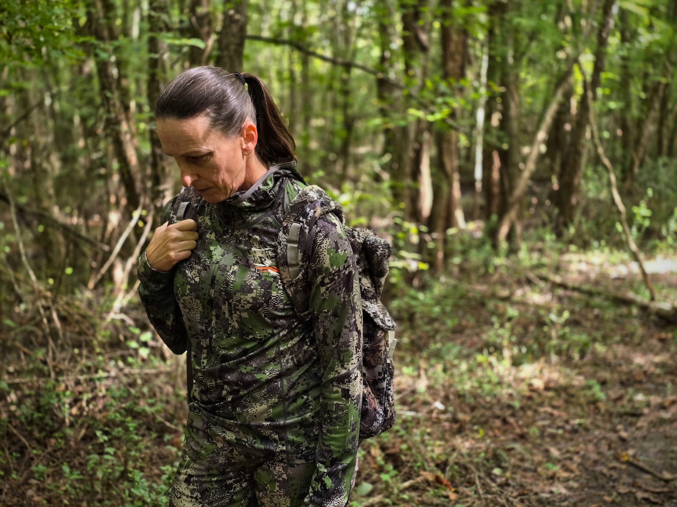 A woman dressed in vibrant green turkey hunting camo walks through the woods wearing a backpack.