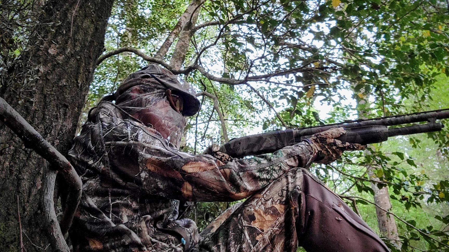 A turkey hunter wearing Realtree camo and a facemask holds a shotgun and leans against a tree trunk.