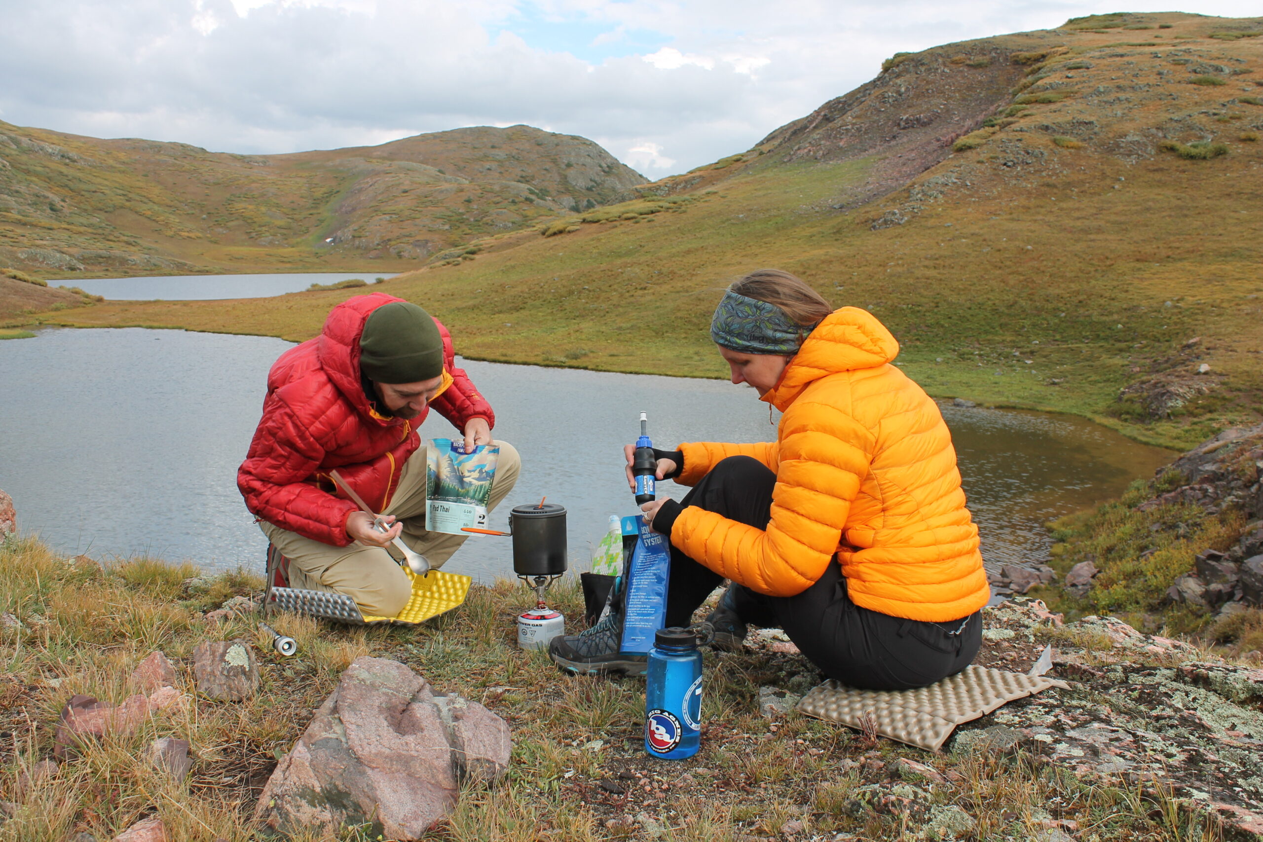 Two campers sit on ground pads while cooking a meal on a camp stove. 