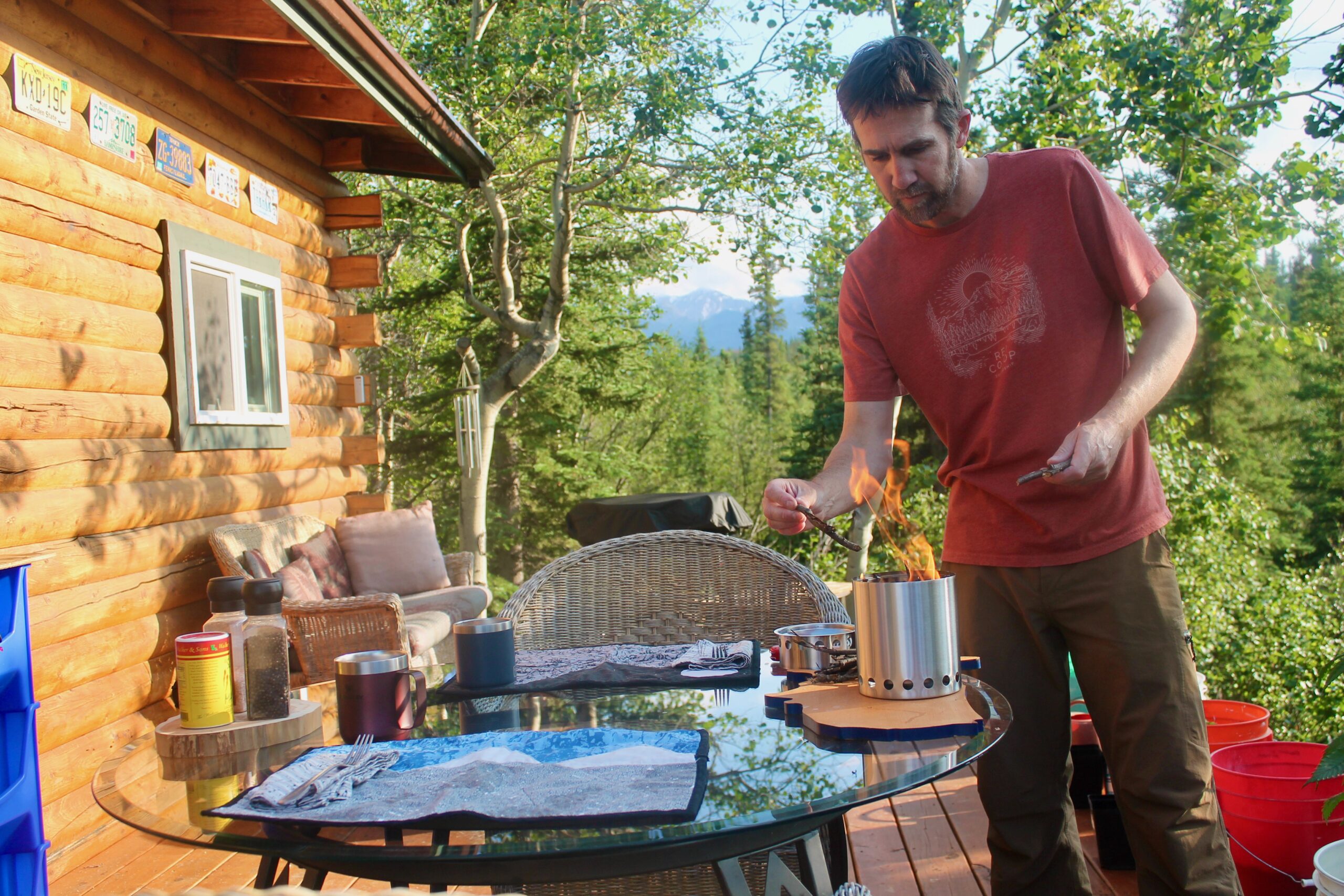 A man feeds twigs to a camping stove on the deck of a cabin. 