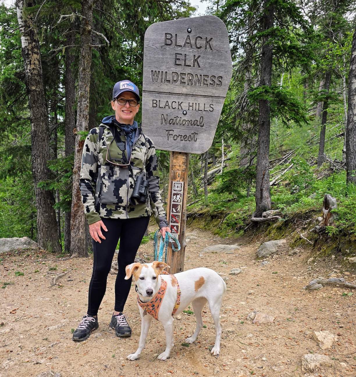 A female hiker in leggings with a dog stand on a trail next to a Black Elk Wilderness sign.