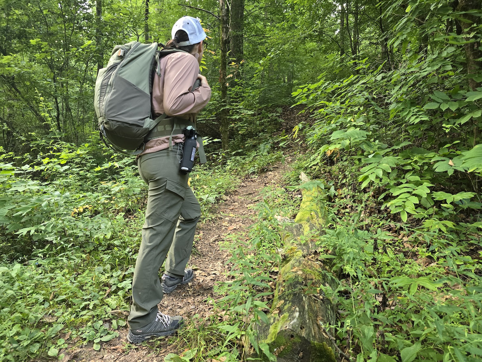 A woman with a backpack and a bear spray canister hikes up a mountain trail.